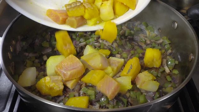 Normal speed followed by slow motion: Close POV overhead shot of chunks of mixed raw fish (cod, smoked haddock and pink salmon) being tipped into a frying pan with vegetables on an oven hob.