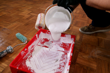 Construction worker and handyman doing finish renovation at apartment. Professional painter with black gloves pours white color from bucket into red bowl for paint mixing. Home renovation concept.