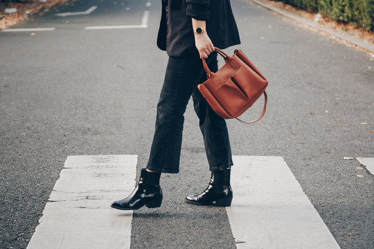 fashion blogger autumn 2019 outfit details. fashionable woman wearing black jeans, black ankle shoes a brown tan trendy leather handbag. crossing the street. detail of a perfect fall fashion outfit. 