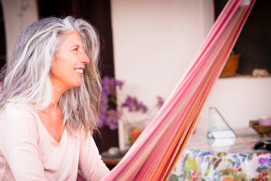 Cheerful beautiful aged adult woman with long grey and white hair - relaxing on an hammock in the garden - people and outdoor leisure activity at home
