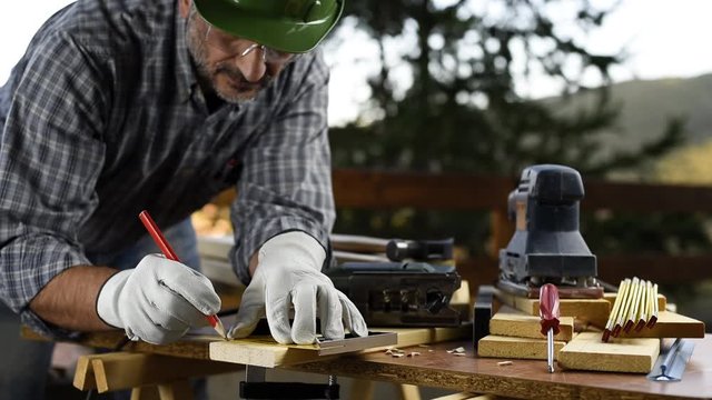 Adult carpenter craftsman wearing helmet and leather protective gloves, with a pencil and the carpenter's square trace the cutting line on a wooden table. Construction industry, housework do it yourse
