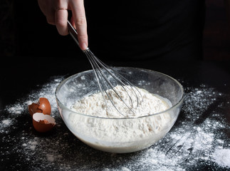 The cook mixes the ingredients with a whisk in a glass bowl with milk and eggs and flour to prepare a delicious meal. Pancake Recipe. Dark background.
