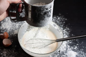 The cook sprinkles flour through a sieve into a glass bowl with milk and eggs to prepare a delicious meal. Pancake Recipe. Dark background.