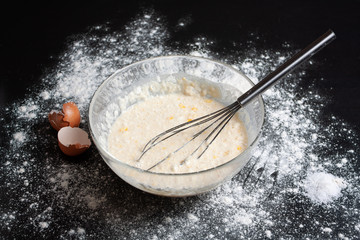 On a dark background is a glass bowl with ingredients for pancakes, eggs, flour, salt, milk. Traditional cuisine. Tasty food. Pancake Recipe. Dark background.