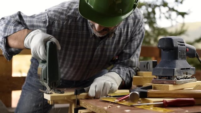 Adult carpenter craftsman wearing helmet and leather protective gloves, with electric saw working on cutting a wooden table. Construction industry, housework do it yourself. Footage.