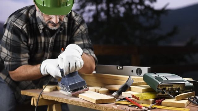 Adult carpenter craftsman wearing helmet and leather protective gloves, with the electric sander smoothes a wooden table. Construction industry, housework do it yourself. Safety at work.