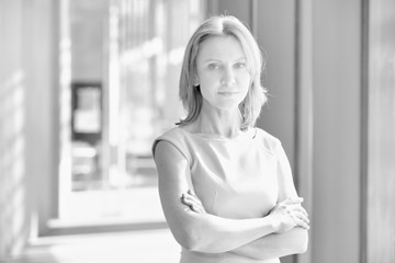 Black and white photo of mature businesswoman standing with arms crossed in office 