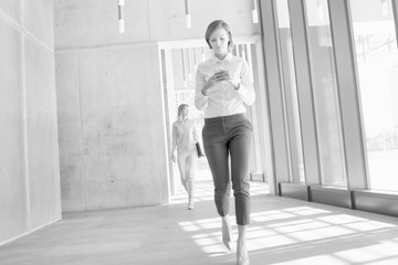 Black and white photo of businesswoman using smartphone while walking in office hall