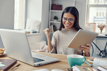 Beautiful young woman in casual clothing using laptop and cheering while working indoors