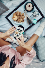 Close up top view of young woman in pajamas photographing flat lay of breakfast while resting in bed at home