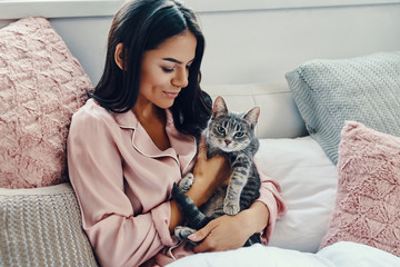 Beautiful young woman in pajamas smiling and embracing domestic cat while resting in bed at home