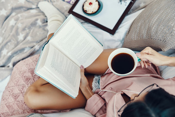 Top view of beautiful young woman in pajamas reading book and enjoying morning coffee while resting...