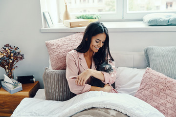 Beautiful young woman in pajamas smiling and embracing domestic cat while resting in bed at home