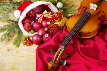 Violin on burgundy drapery, Santa Claus hat with colored balls and gifts on a wooden table.