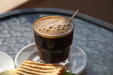 Close-up of a glass of black coffee with foam, a sandwich on a white plate.A light Breakfast on a glass table or a short coffee break with informal communication.Selective focus
