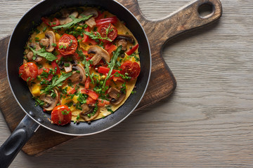 top view of yummy omelet with vegetables and greens in frying pan on wooden board