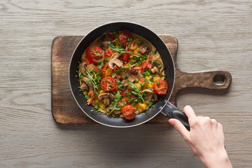 top view of woman cooking omelet with mushrooms, tomatoes and greens on frying pan on wooden board