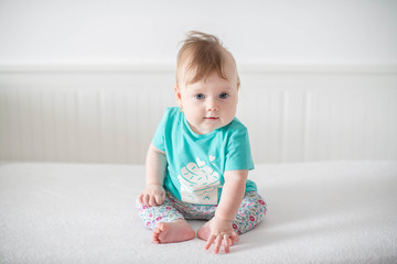 Fototapeta premium Portrait of adorable Caucasian baby girl with blue eyes, looking at the camera calmly, with curiosity, interactivity or inquisitiveness and sitting in a pristine white baby cot while holding her toes