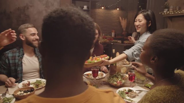 Tracking shot of hospitable Asian woman smiling and serving plate of open sandwiches to cheerful guests during dinner party in her cozy kitchen