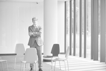 Black and white photo of Confident mature businessman standing with arms crossed amidst chairs in new office