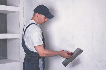 Worker in overalls and cap plastering a wall with finishing putty using a putty knife. Repair work and construction concept