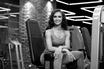 Obraz na płótnie Canvas Sexy fitness brunette woman is sitting in training apparatus for legs in gym BW