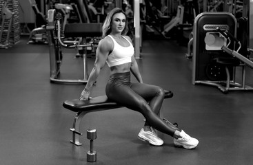 Fototapeta na wymiar Blonde bodybuilder woman is sitting with dumbbell in the gym on the bench BW
