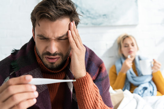 sick man with headache and fever looking at thermometer in bedroom with woman behind