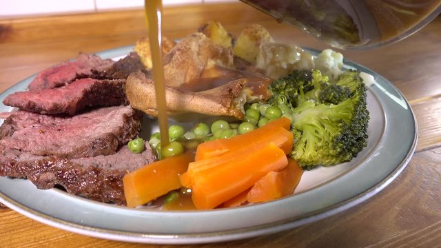 Close POV circular dolly shot of steaming thick gravy being poured all over a Sunday dinner plate on a pine kitchen worktop, of traditional roast beef, vegetables and Yorkshire pudding.