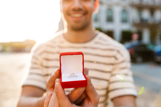 Image of cheerful masculine man smiling and holding gift box with ring