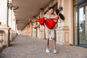 Image of young unshaven man carrying happy woman in hands and smiling
