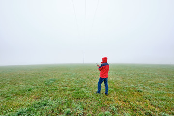 Rear view of man in red jacket standing on a green meadow and looking on his smartphone in front of...