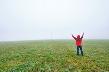 Rear view of man in red jacket standing on a green meadow and raising his arms in joy in front of a foggy nowhere landscape. Seen in October in Germany, Bavaria near Oedenberg.