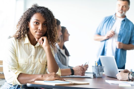 Beautiful cheerful African woman looking at camera with smile while sitting at the office table with her coworkers.