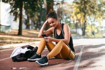 Young fitness woman in park. Athletic woman sitting on  running track and using phone.
