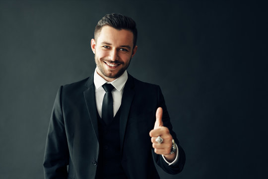 Smiling young man in suit showing thumbs up sign on black background