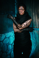 man with tree makeup for halloween party on his face and hands poses at night against a wooden gate in cyan color smoke