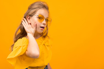 beautiful little blonde in yellow glasses eavesdrops, a girl on a yellow background holds a palm by...