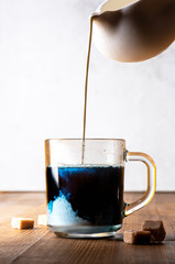 Blue matcha with cream. Pouring cream from white creamer into hot blue matcha in a glass cup on a...