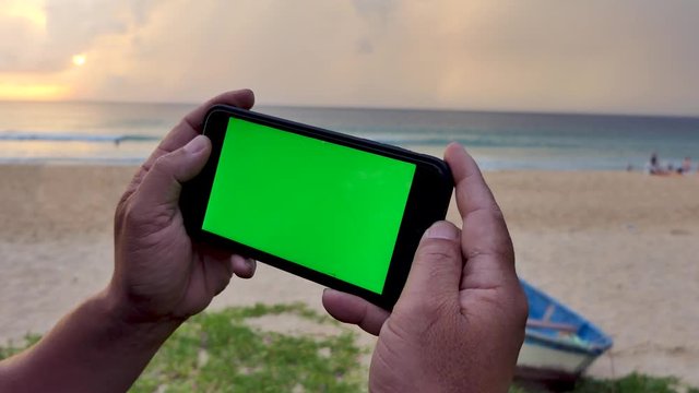 The man's hand with the green screen phone on the beach at sunset.