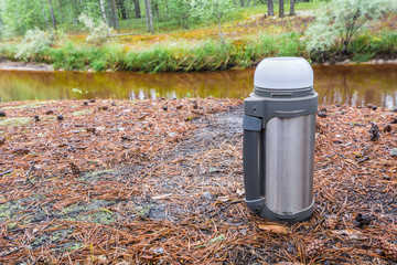 Hot tea thermos stands on ground with pine needles in forest against taiga river.