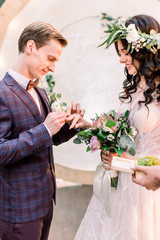 Wedding couple exchange rings on wedding ceremony outdoors. Bride with a bouquet of flowers and greenery and groom in elegant suit with arch on background, rustic style