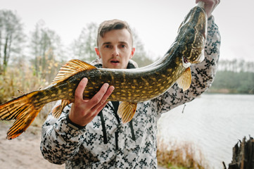 Amazed cheerful young fisherman hold a big fish pike near the boat on a background of lake and...