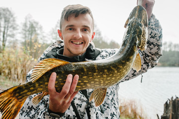 Happy cheerful young fisherman hold a big fish pike on a background of lake and nature. Fishing...