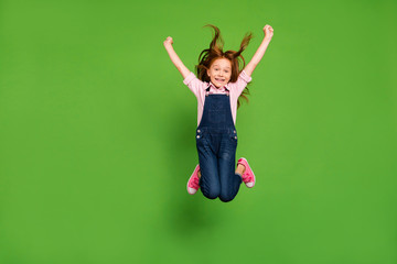 Full body photo of pretty little schoolchild jumping high rejoicing summer holidays hair flying warm wind wear casual denim overall pink shirt isolated green background