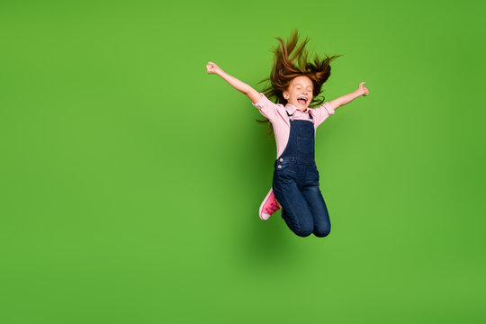 Full length photo of cheerful pretty little schoolchild jumping high rejoicing summer holidays hair flying wear casual denim overall pink shirt isolated green background