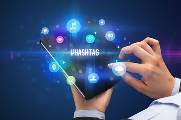 Businessman holding a foldable smartphone with HASHTAG inscription, social media concept
