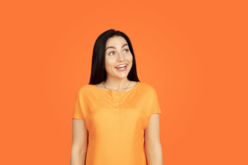 Obraz premium Caucasian young woman's portrait on orange studio background. Beautiful female brunette model in shirt. Concept of human emotions, facial expression, sales, ad. Copyspace. Looks dreamful, smiling.