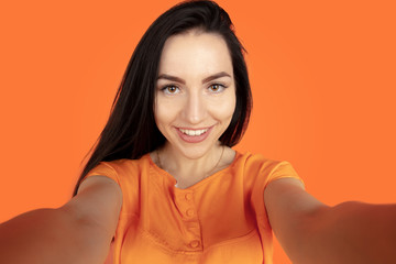 Fototapeta premium Caucasian young woman's portrait on orange studio background. Beautiful female brunette model in shirt. Concept of human emotions, facial expression, sales, ad. Copyspace. Making selfie and smiling.