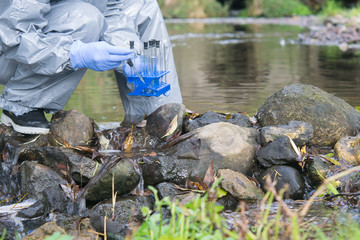 a specialist in a protective suit and rubber boots draws water from a river into various test tubes...